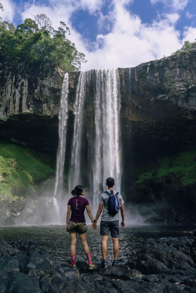 A couple are pictured, standing at the edge of the water at the bottom of a waterfall centered in the frame directly in front of them. They hold hands while dressed in hiking clothing, including backpacks.