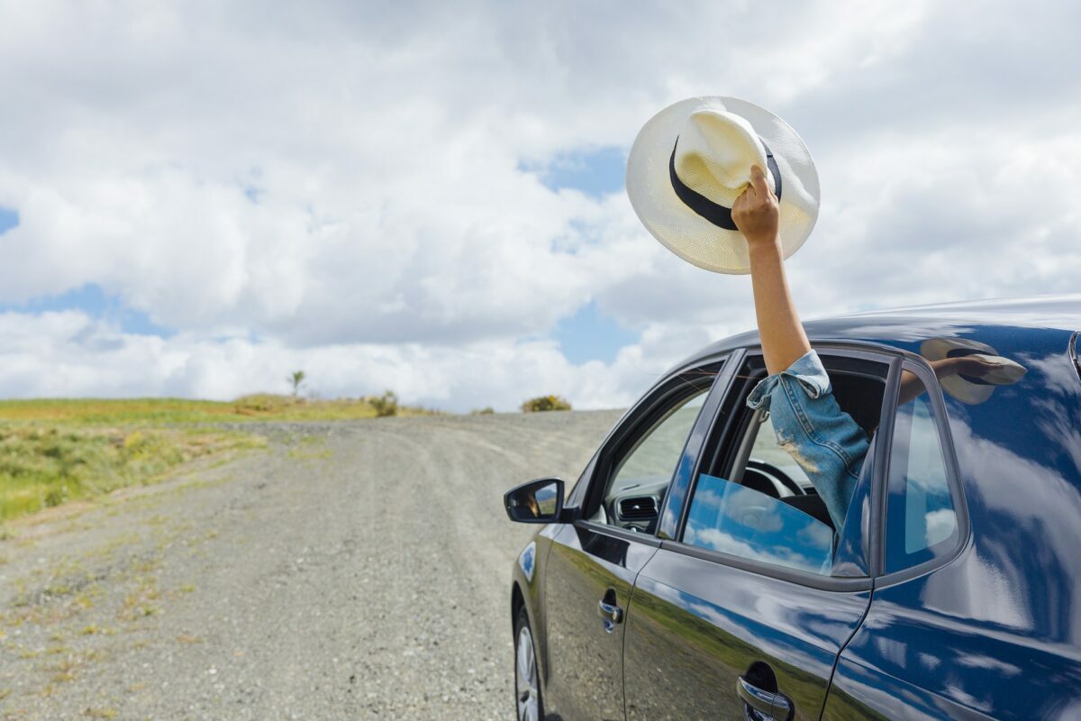 A dark, 4-door sedan tours down a dirt road on a somewhat bright yet cloud-covered day. An arm of the left-rear passenger can be seen joyfully waving a wide-brimmed white hat with black band in the air out of their window.