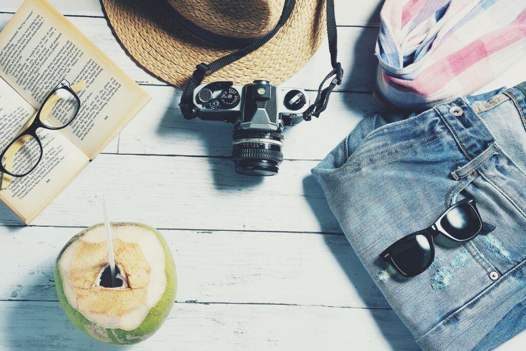 A coconut with a hole at the top and a straw sticking out of it, a camera, jeans, sunglasses, a map, and a wide-brimmed straw hat can be seen in this picture, potentially eluding to an upcoming vacation.