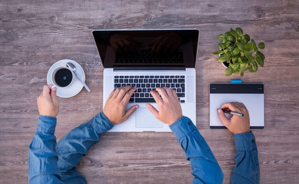 A picture of a man in a blue dress shirt with four arms. He's typing on a MacBook, Writing on a digital tablet with a tablet stylus or pen, and reaching for his cup of coffee, all seen by us from an overhead view.