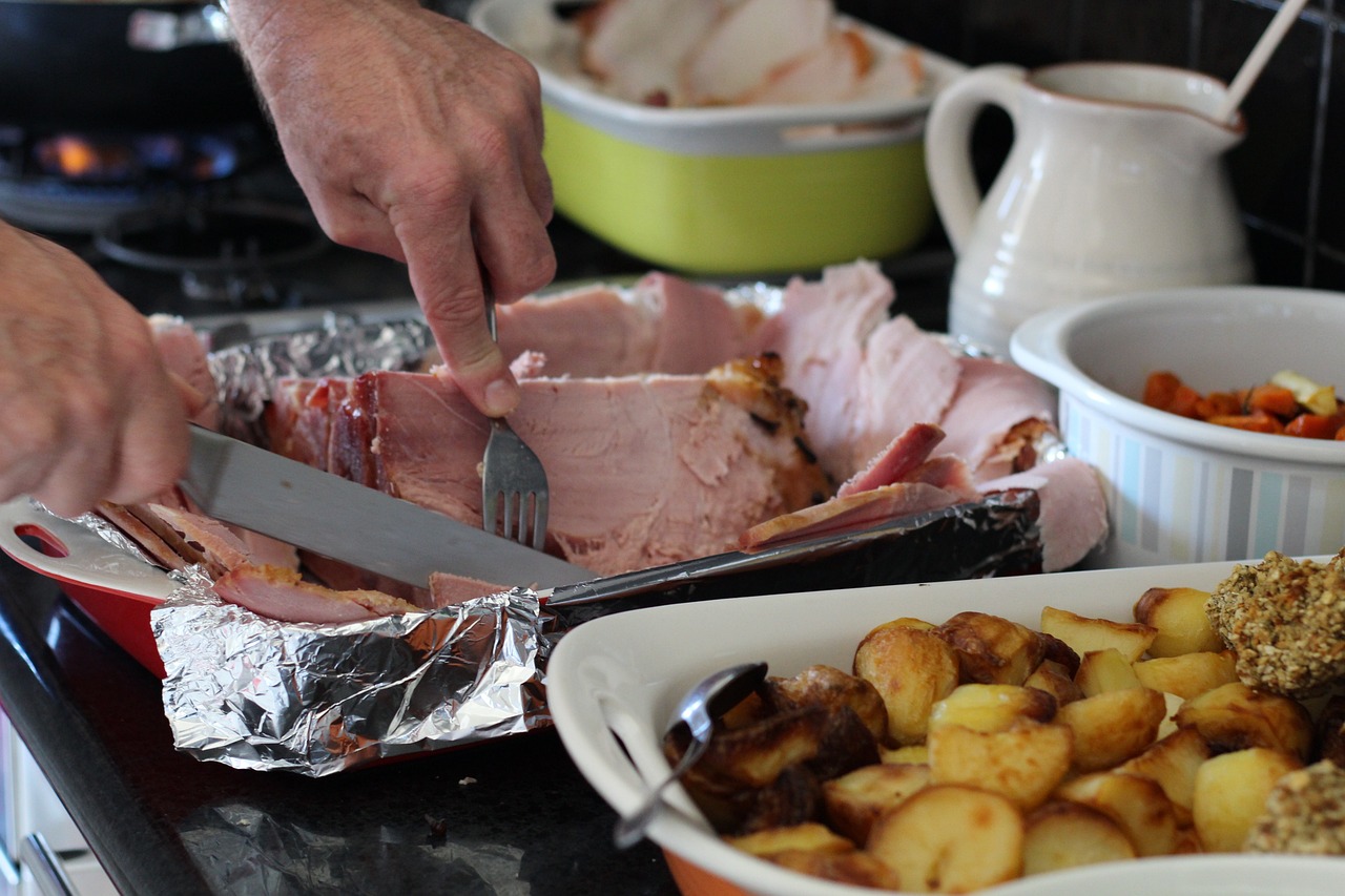A Ham Dinner laid out on the table with a large roast of ham being sliced inside the foil-lined roaster, potatoes and other veg on the side, and more.