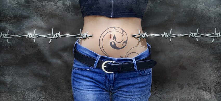 IBS is the theme here, with a picture of barbed-wire running across the picture, passing through the left and right sides of a woman's stomach. Around her bellybutton is a black-ink tattoo. She's wearing a bellyshirt, and jeans with a black leather belt.