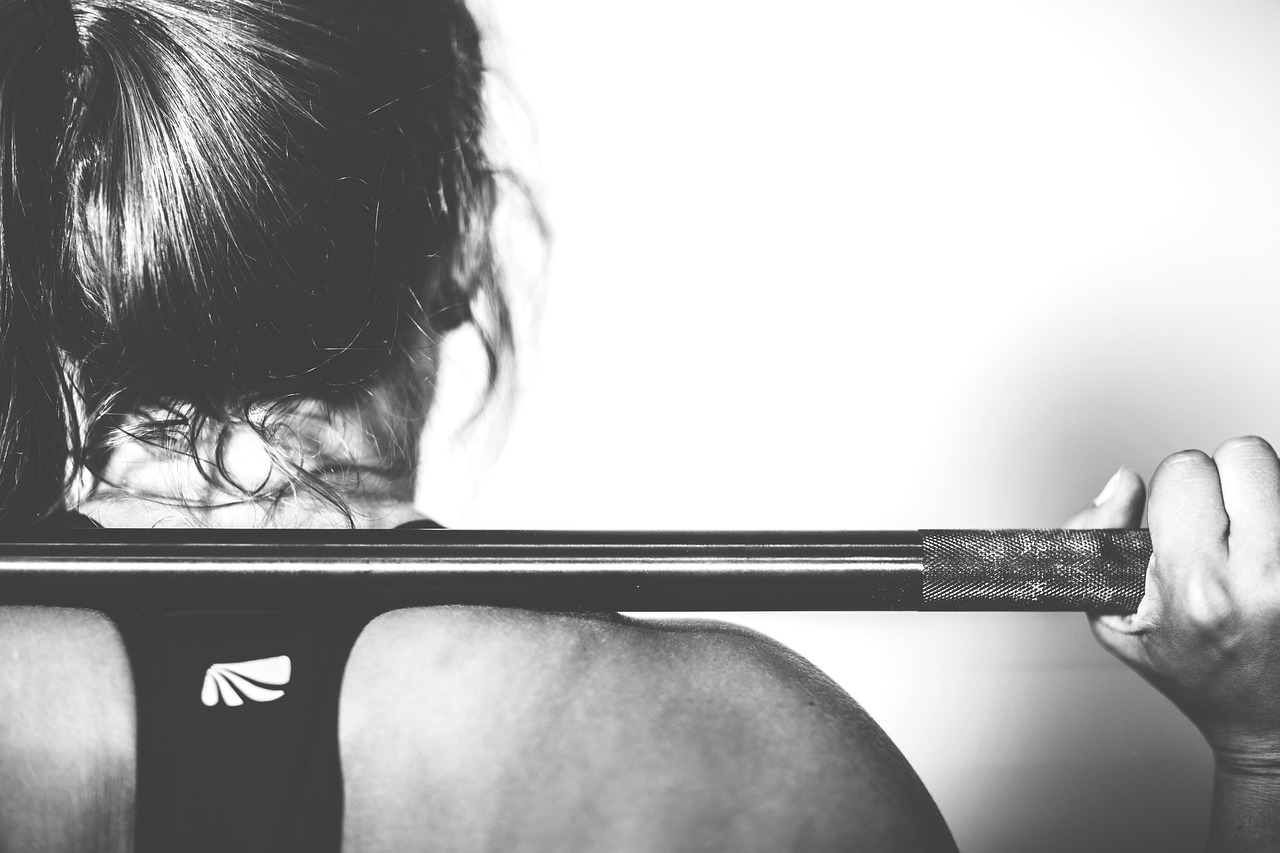 An athletic woman, hair up in a ponytail or bun, black sport-support tank top on. A weight bar is across her shoulders, hands placed to stabilize it as she exercises.