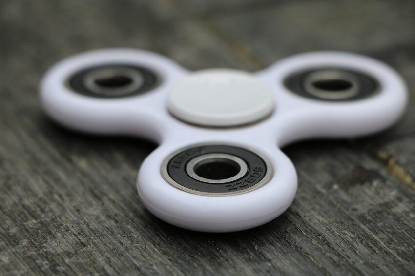 A fidget-spinner is pictured on a wooden surface that appears to have been stained in a dark grey stain. The fidget-spinner is white, with dark inner circles.