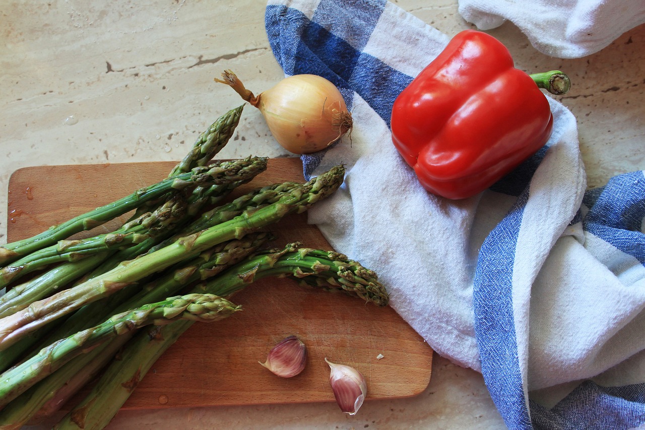 Some vegetables (some of which would be considered prebiotic in nature) are pictured on a wooden cutting board. We see asparagus, garlic, chalets, and a red pepper sitting on a dish towel sporting larger-print white and blue squares.