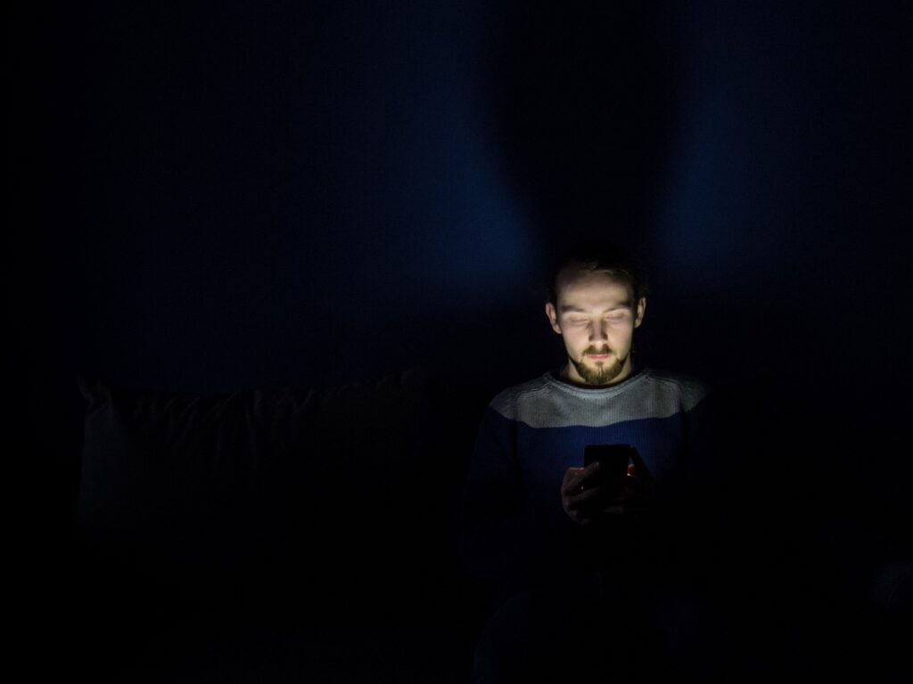 A man in the darkness, scrolling on his mobile device while in bed. Only his upper-body and face can be seen by the light of the device's screen, as he leans with his head and shoulders propped up against his headboard.