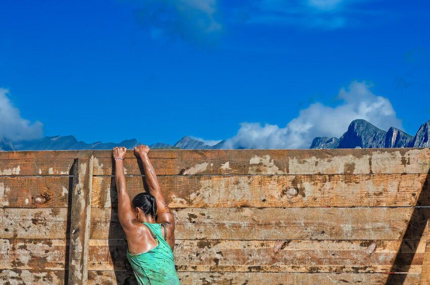 An athletic-looking woman in a green tank top hangs from an obstacle course wall that it seems she's attempting to get to the other side of. It would suggest she's participating in a race of some sort.