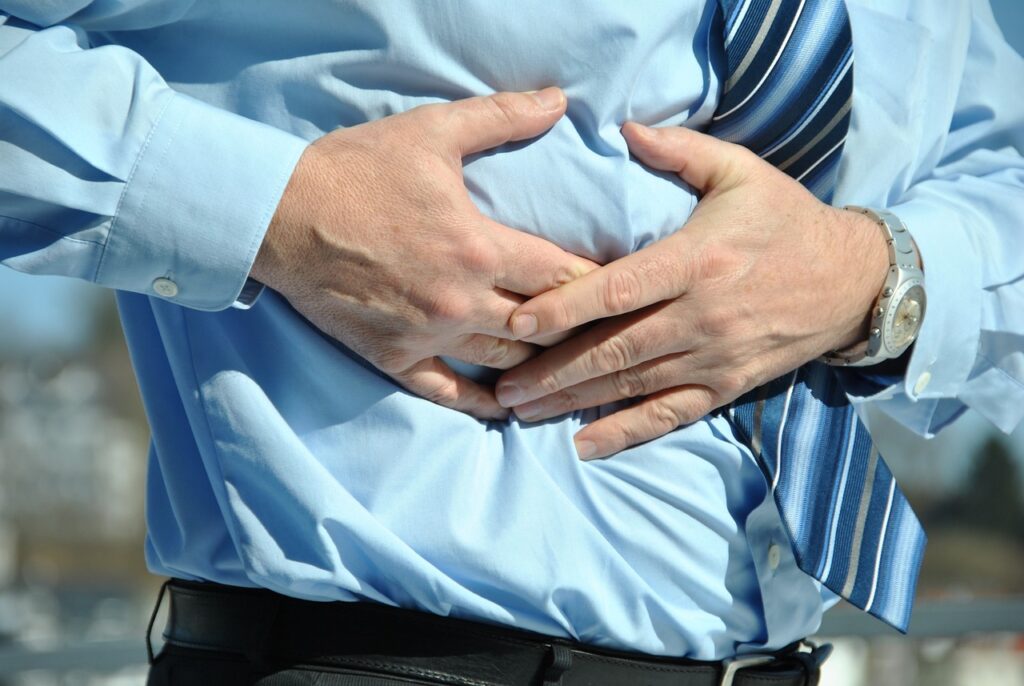 A man's torso is shown with him holding his hands to his side as if palpating the area, or in pain. He's dressed in a blue dress shirt, a blue diagonal-striped tie, and a silver watch on his left wrist.