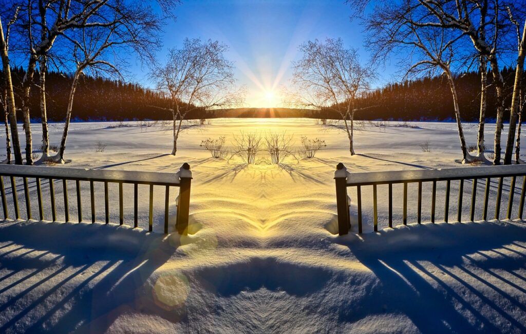 An early-morning sunrise coming up between two mountain peaks off in the distance. The view is from a back deck, snow down through the back, across a snowy field. Backyard fence in the foreground. 