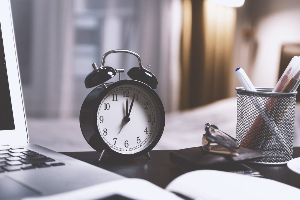 A dual-bell alarm clock sits at the back of a desk. Part of a computer keyboard, an open book, and a pencil cup are all seen around the desk as well.