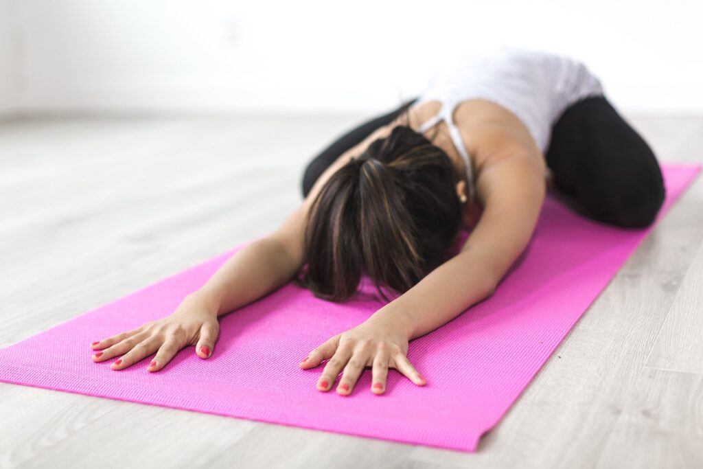 A woman dressed in a white tank top and black yoga pants stretches out, face-down with her hands stretched to the top of her bright-pink yoga mat.