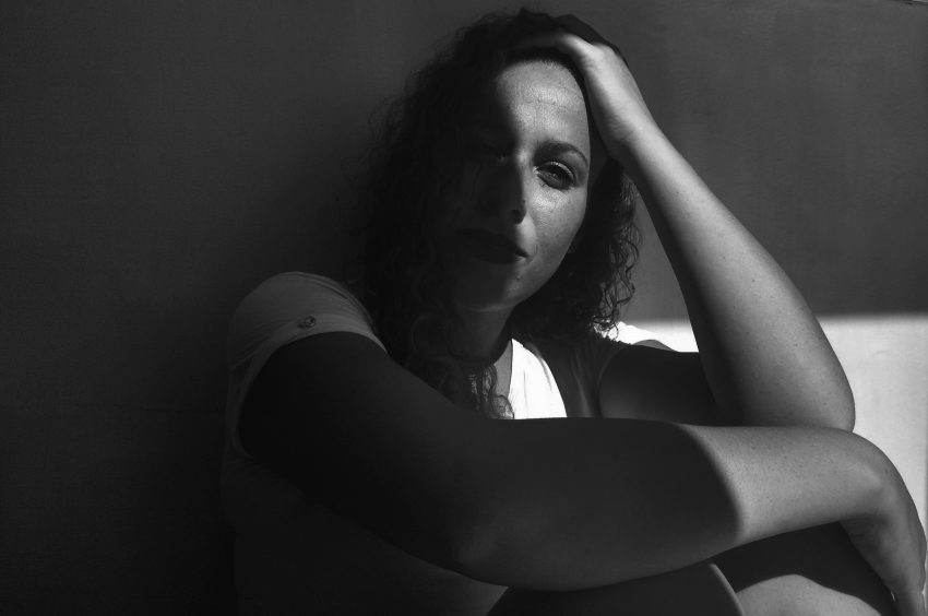 A woman sits partly in a shadow. White t-shirt, head in one hand. Clearly upset or battling a mental health challenge, she stares at the camera.