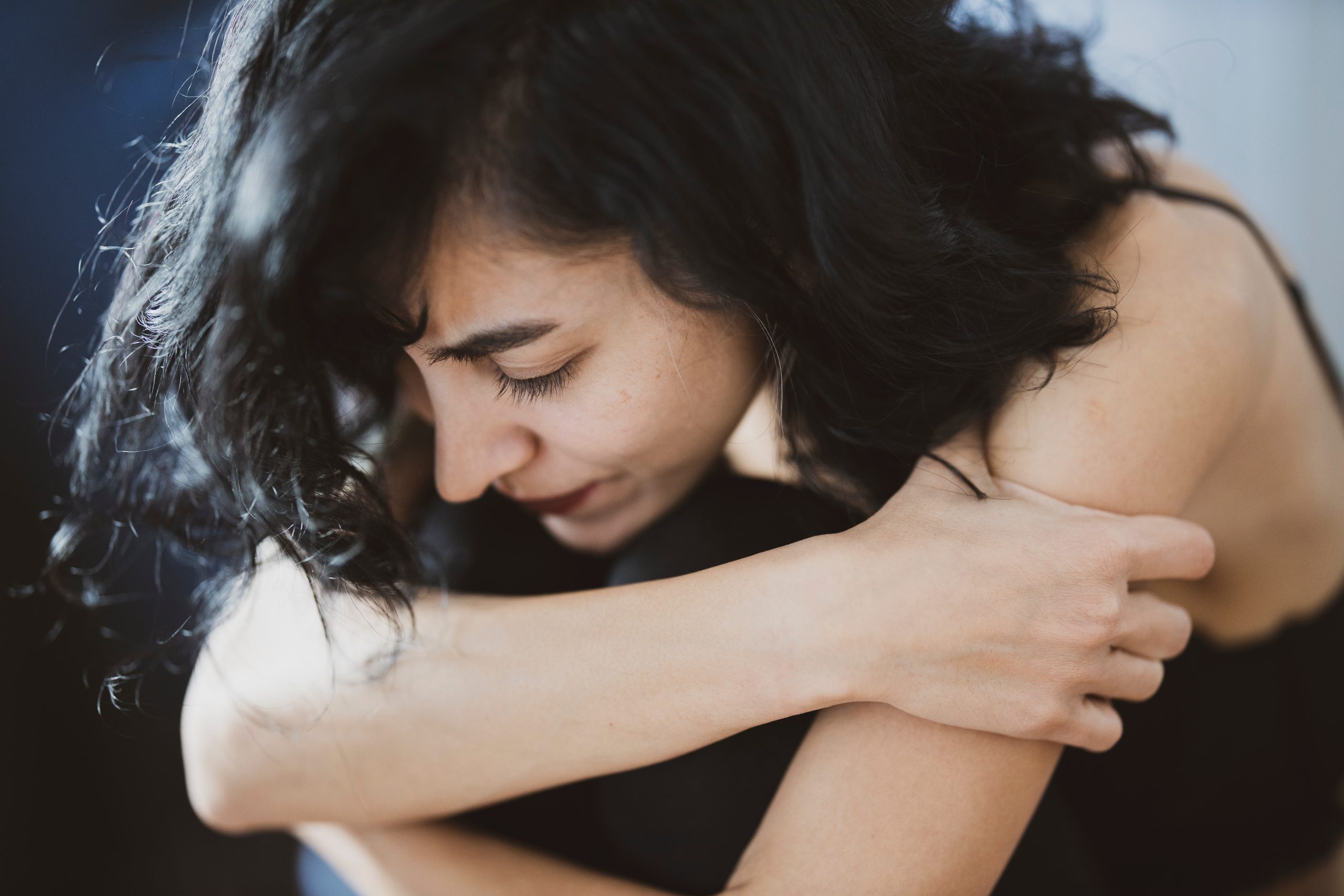 A caucasian woman with a full head of dark, curly and wavy hair is seen with her arms hugging her legs - knees up to her chest, with her chin on her knee. Dressed in a dark tank top and jeans, she is clearly upset or in pain.