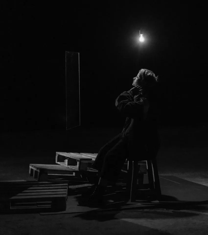 A lone woman with short blonde hair sits in a dimly lit room, surrounded by darkness and wooden pallets, looking up with a sad or anxious expression.